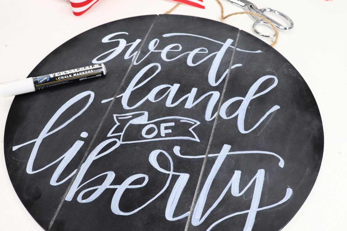 Image contains a round chalkboard sign with the words "sweet land of liberty" written in white chalk marker. The marker sits on top of the sign and there are scissors and ribbon in the background.
