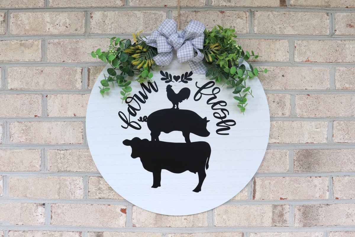 Image contains a white wood circle with a cow, pig, and chicken in black vinyl surrounded by the words "farm fresh." At the top, there is a spray of faux eucalyptus and a grey and white checkered bow.
