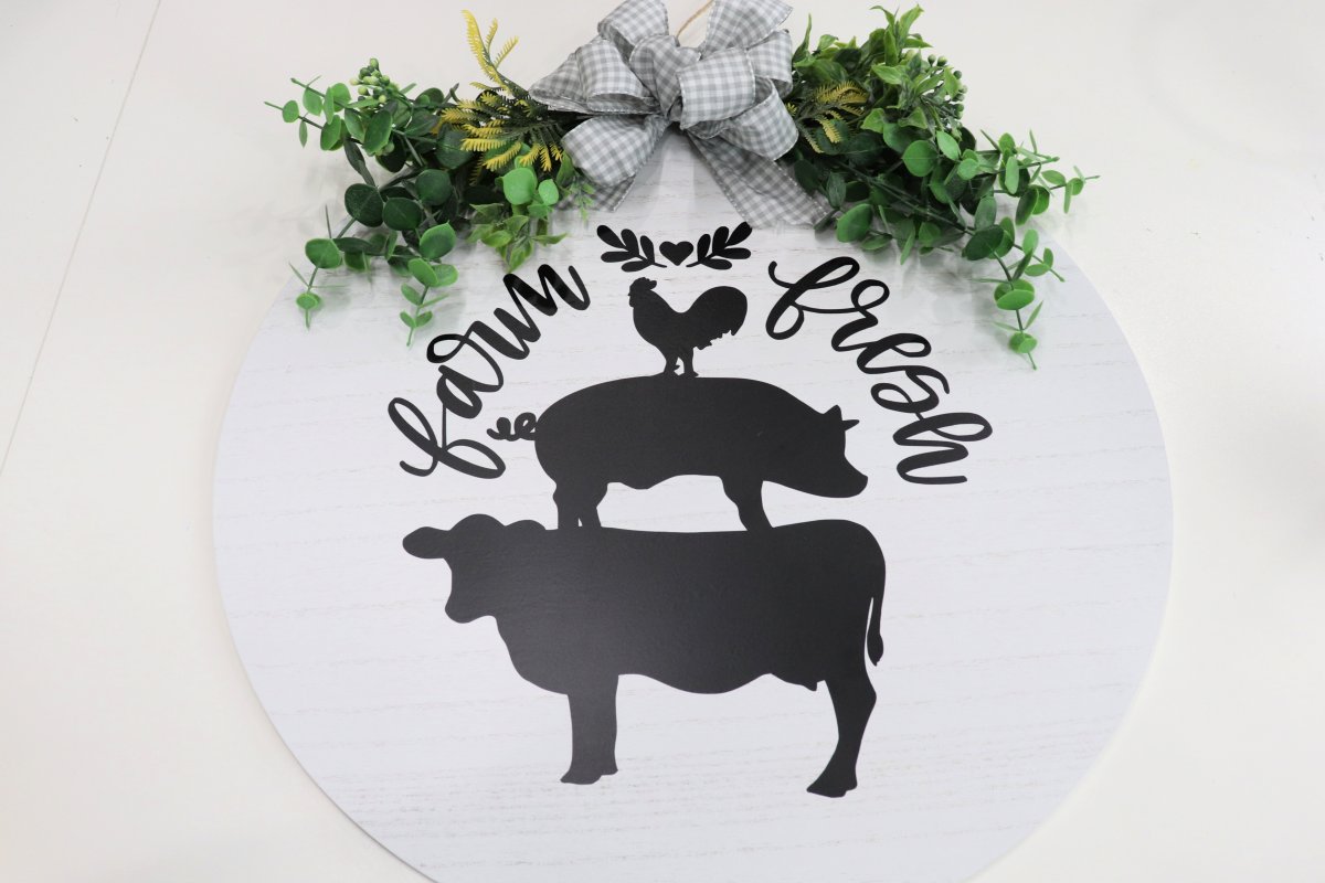 Image contains a white wood circle with a cow, pig, and chicken in black vinyl surrounded by the words "farm fresh." At the top, there is a spray of faux eucalyptus and a grey and white checkered bow.