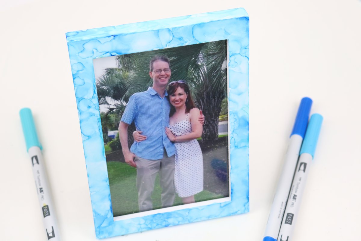 Image contains a photo frame colored in various shades of blue, with a photo of Dan and Amy inside. It sits on a white background surrounded by three blue markers.
