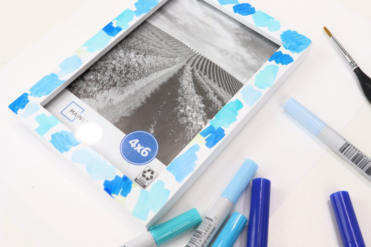Image contains a white photo frame with various shades of blue marker scribbled around it. The frame sits on a white background surrounded by markers and a paintbrush.