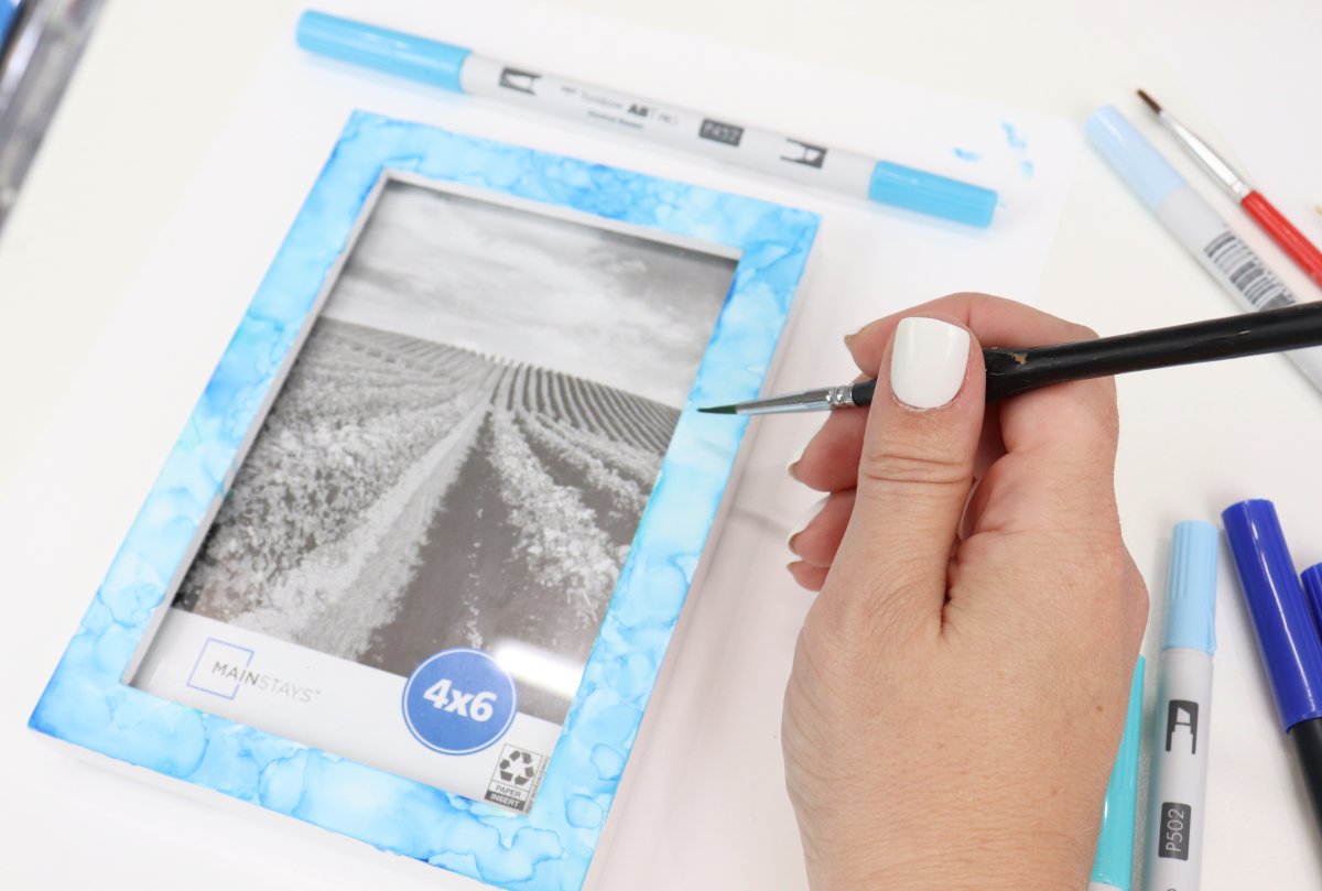 Image contains Amy's hand holding a paintbrush that is blending shades of blue on the edges of a photo frame. The frame sits on a white background surrounded by blue markers and another paintbrush.