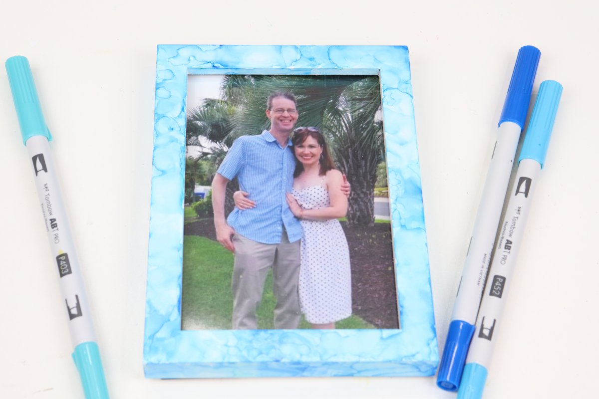Image contains a photo frame decorated in shades of blue and teal with a photo of Amy and her husband inside. It sits on a white background surrounded by three blue markers.
