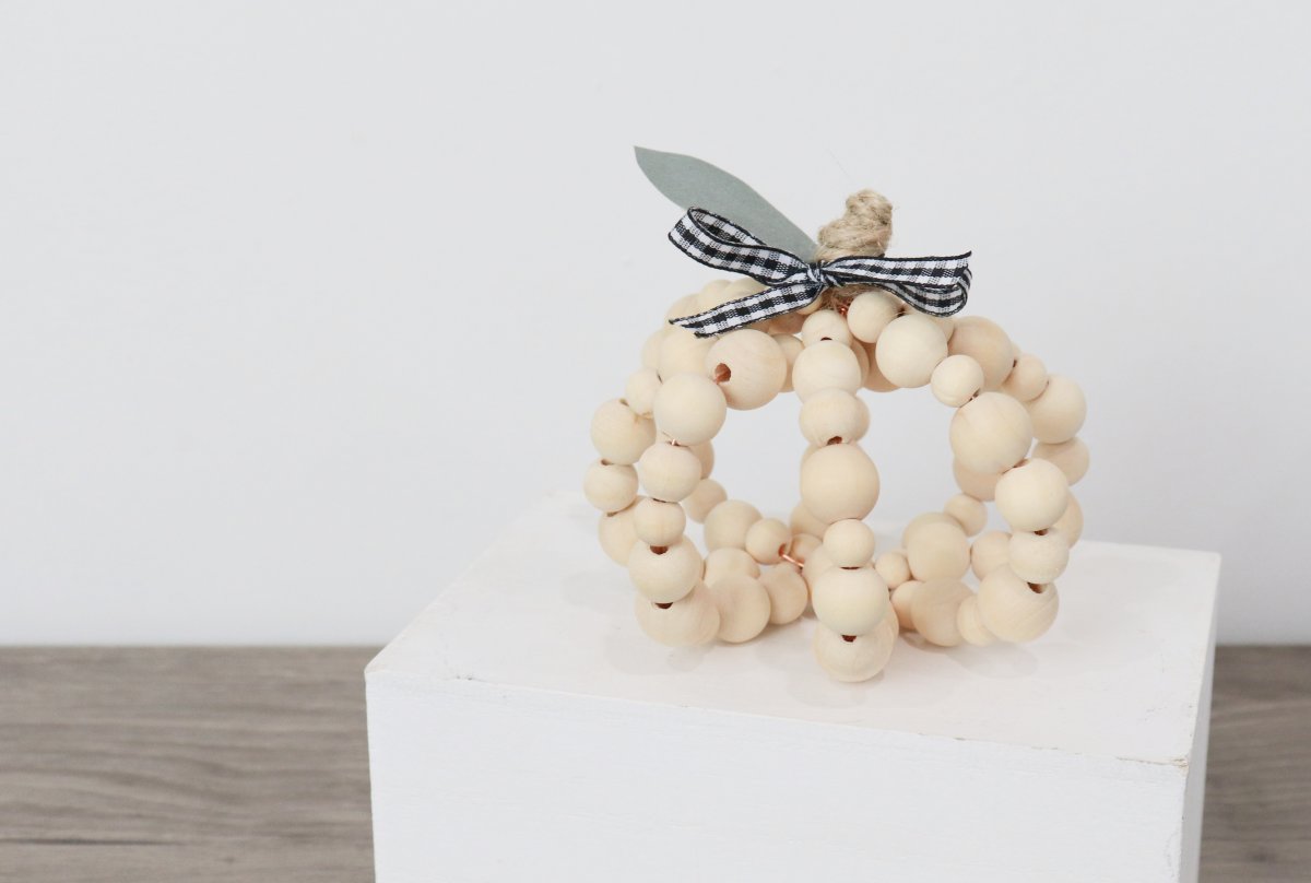 Image contains a decorative pumpkin made of wooden beads with a twine stem, faux leaf, and plaid bow.