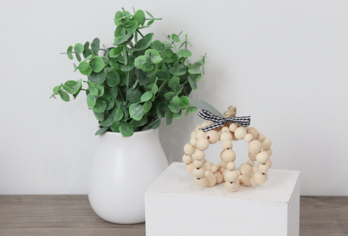 Image contains a pumpkin made of wooden beads sitting on a white box with a vase of eucalyptus in the background.