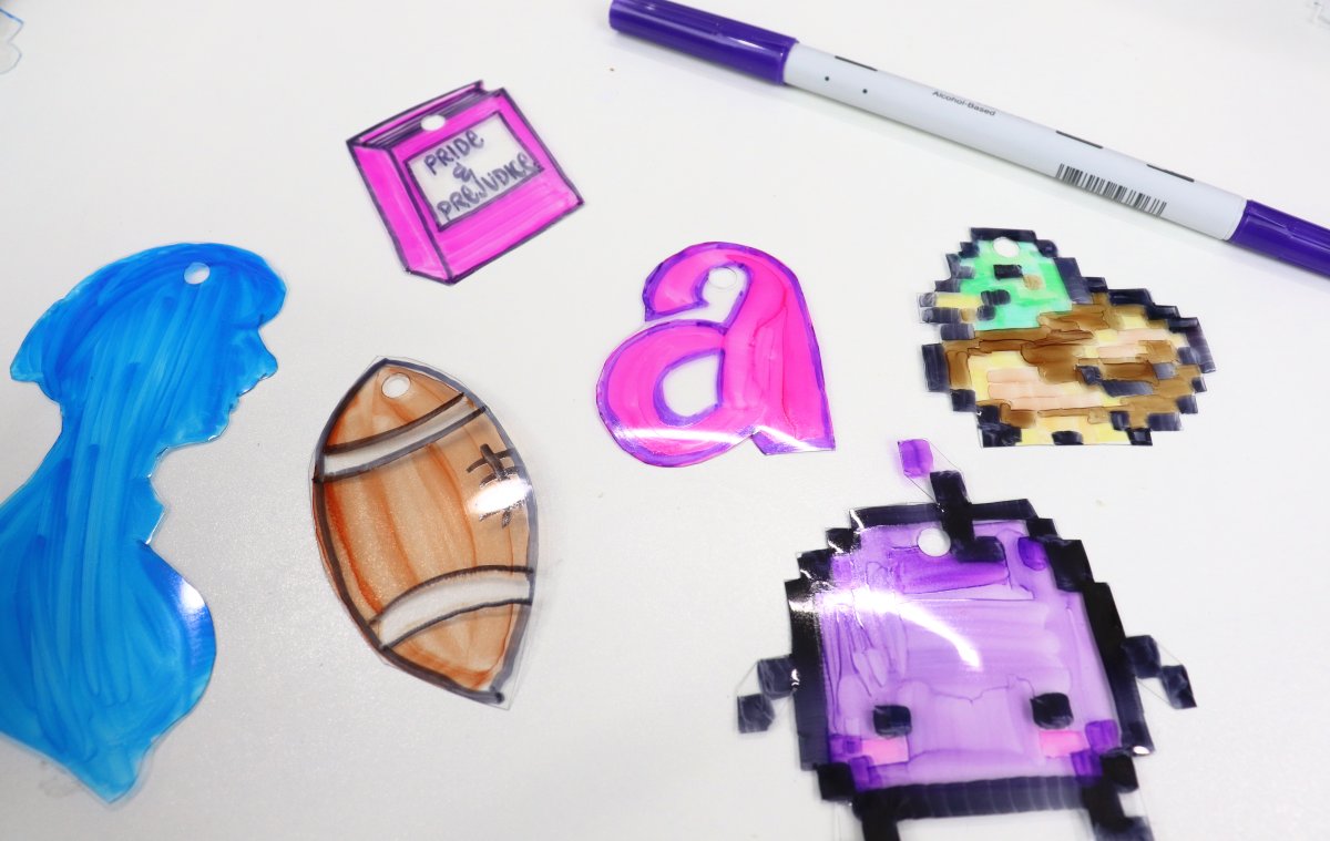 Image contains doodles drawn on shrink plastic and cut out; a blue silhouette of a woman, a pink book, a brown football, a pink monogram "a", a green and brown duck, and a purple Junimo.
