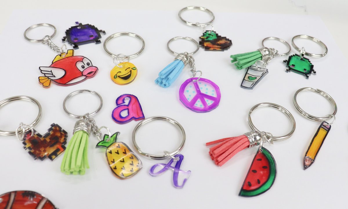 Image contains a colorful assortment of keychains, each with a hand-drawn charm made from shrink plastic. Keychains include a chicken, a pineapple, the letter 'a', an emoji face, a duck, a peace sign, a coffee cup, a watermelon, and a pencil.