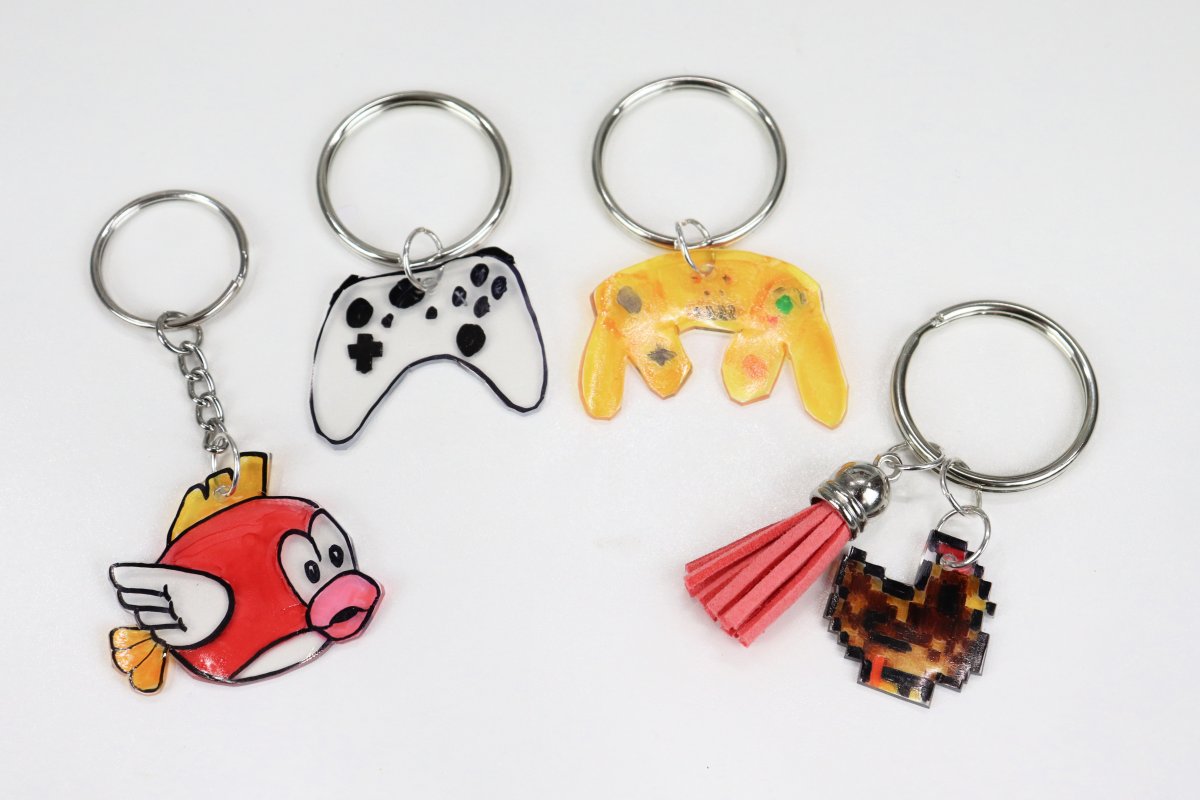 Image contains four keychains; one is the red fish from Super Mario Brothers, one is a white video game controller, one is a yellow video game controller, and one is a brown chicken with a pink tassel.