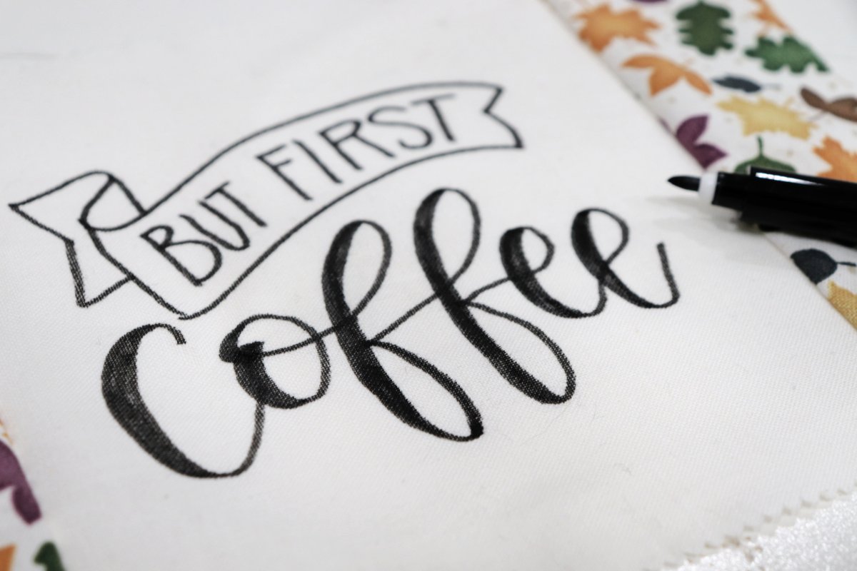Image contains a closeup of a white fabric square with black lettering that reads, "but first, coffee." A black fabric marker sits nearby.