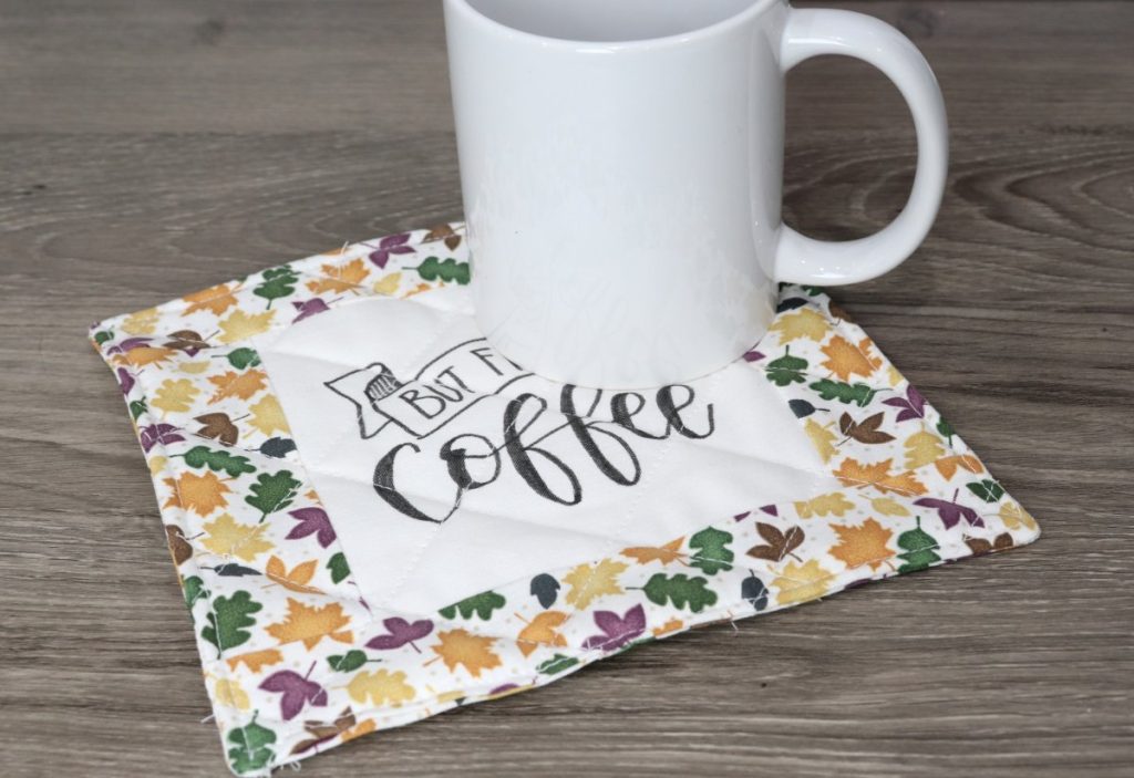Image contains a quilted mug coaster with multicolored autumn leaves under a white mug on a wooden background.