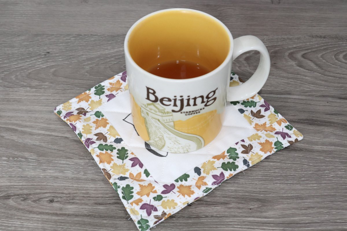 Image contains a quilted coaster with autumn leaf print fabric, and a large coffee mug sitting on top.