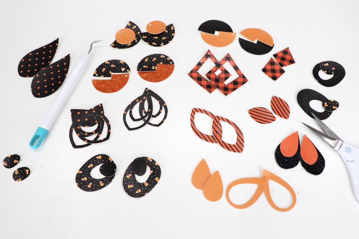 Image contains orange and black faux leather cut into shapes for a dozen pairs of earrings, along with a Cricut weeding tool and a pair of scissors.