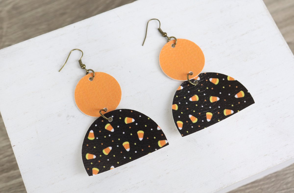 Image contains a pair of faux leather dangle earrings made from an orange circle on top of a larger black and candy corn patterned half circle.