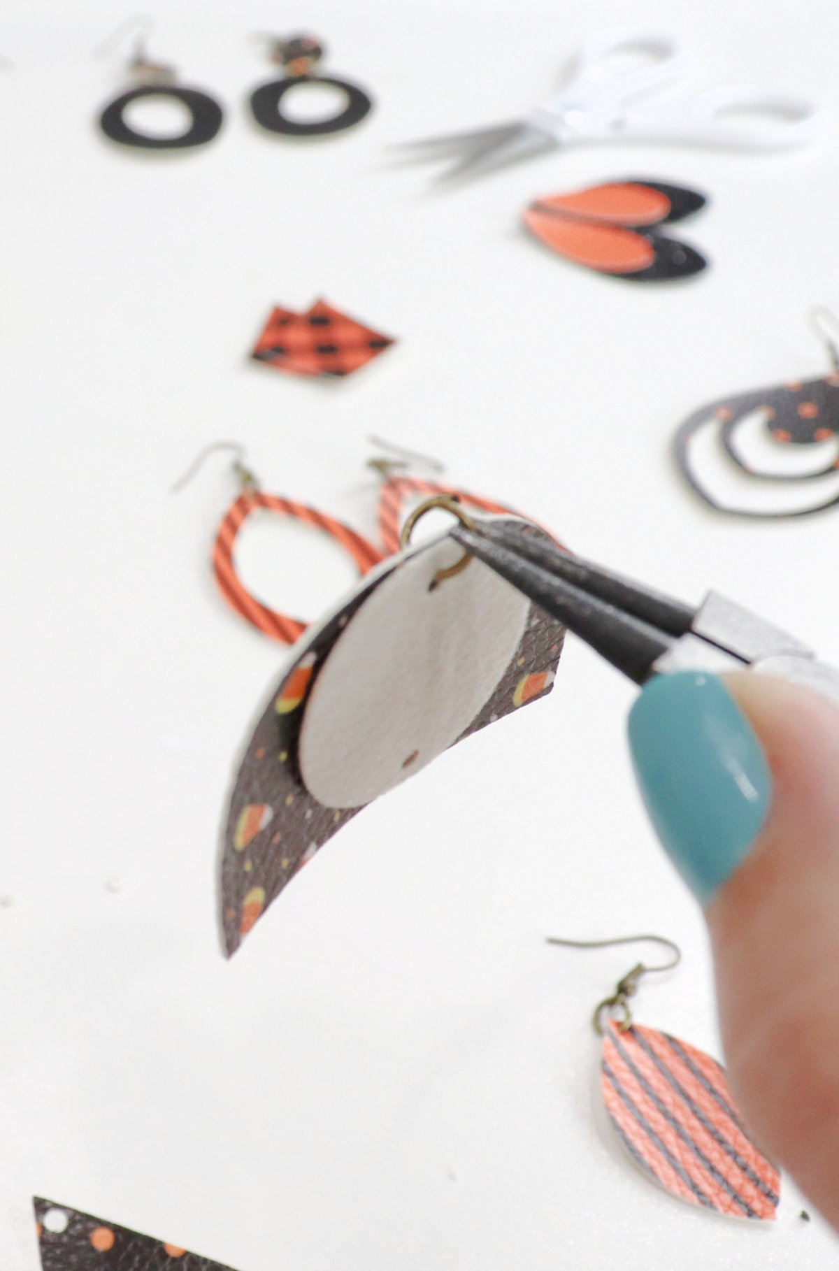 Image shows Amy's hand with a pair of needle nose pliers attaching two pieces of cut faux leather with a jump ring. In the background are multiple pairs of orange and black earrings and a pair of white-handled scissors.