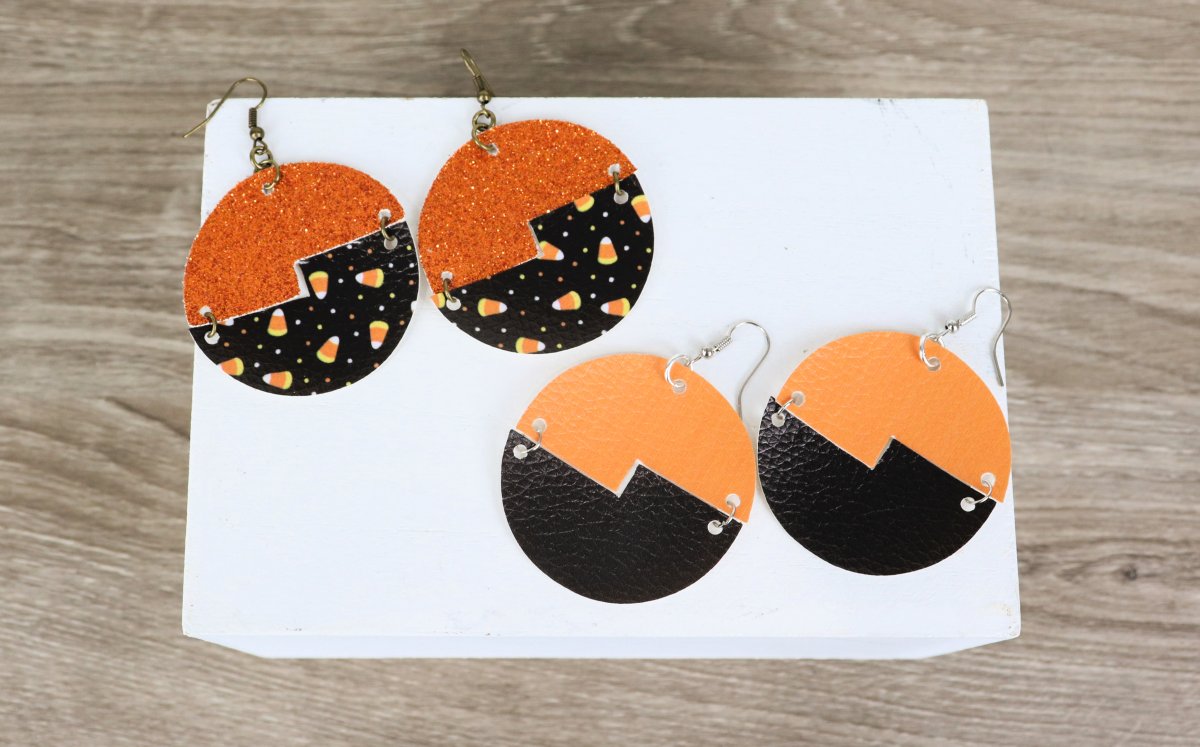 Image contains two pairs of faux leather earrings. They are large circles with the top half cut from one orange vinyl and the bottom from black.