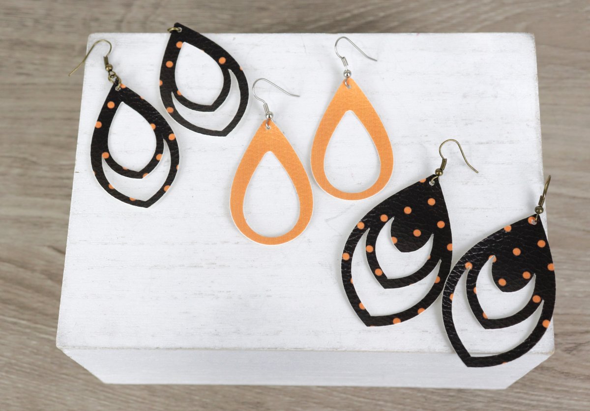 Image contains two pairs of black and orange polka dot dangle earrings and one pair of orange dangle earrings, all teardrop shapes made from faux leather.