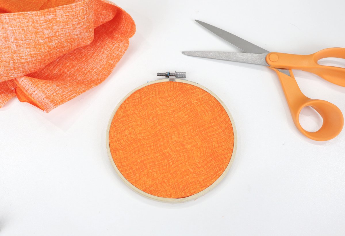 Image contains orange fabric in an embroidery hoop, with orange fabric and orange handled scissors off to the sides.