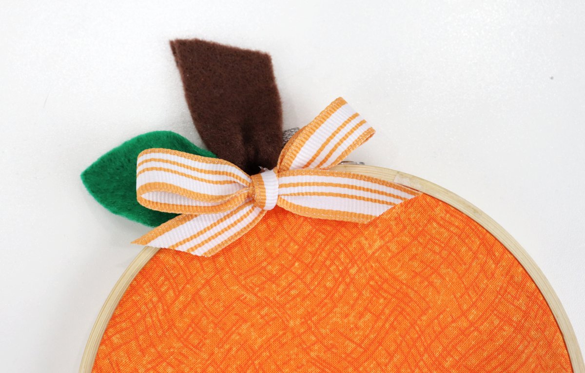 Image contains a close-up of the embroidery hoop pumpkin craft featuring the brown felt stem, green felt leaf, and orange and white ribbon bow.