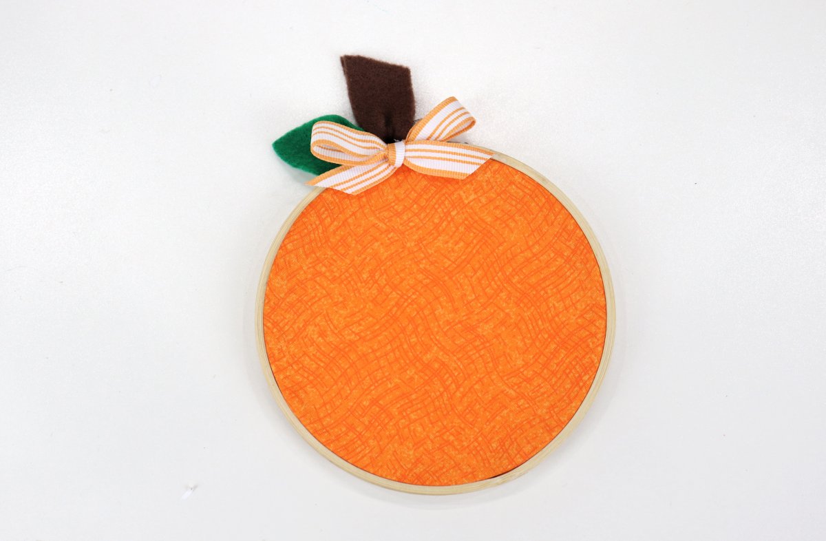 Image contains an embroidery hoop with orange fabric inside, a brown felt stem, a green felt leaf, and a striped orange and white bow on a white background.