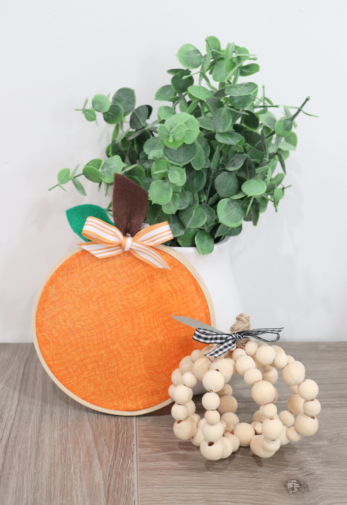 Image contains a pumpkin made from wooden beads, an orange pumpkin made from fabric in an embroidery hoop, and a white vase with faux eucalyptus.