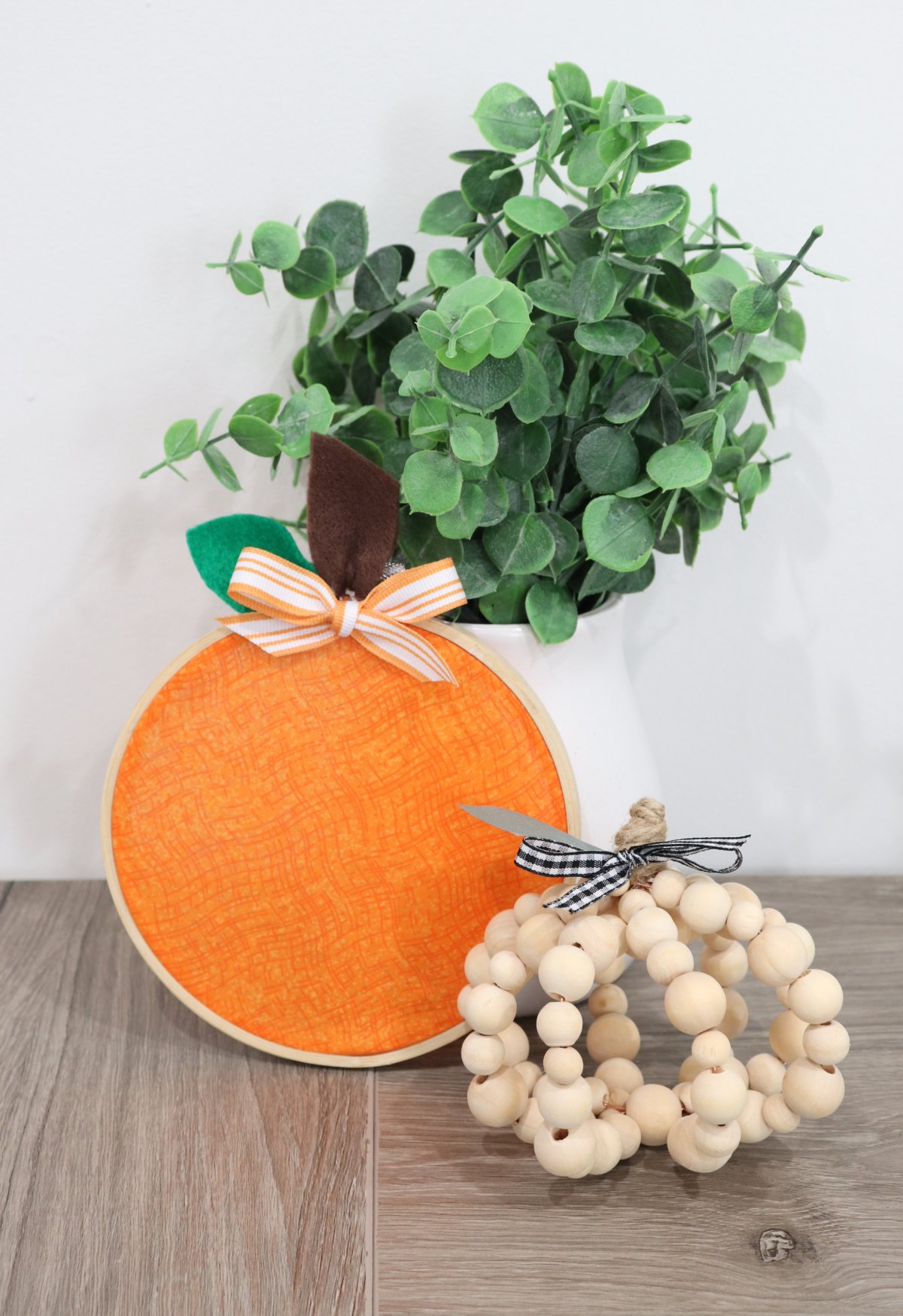 Image contains a pumpkin made from wooden beads, an orange pumpkin made from fabric in an embroidery hoop, and a white vase with faux eucalyptus.