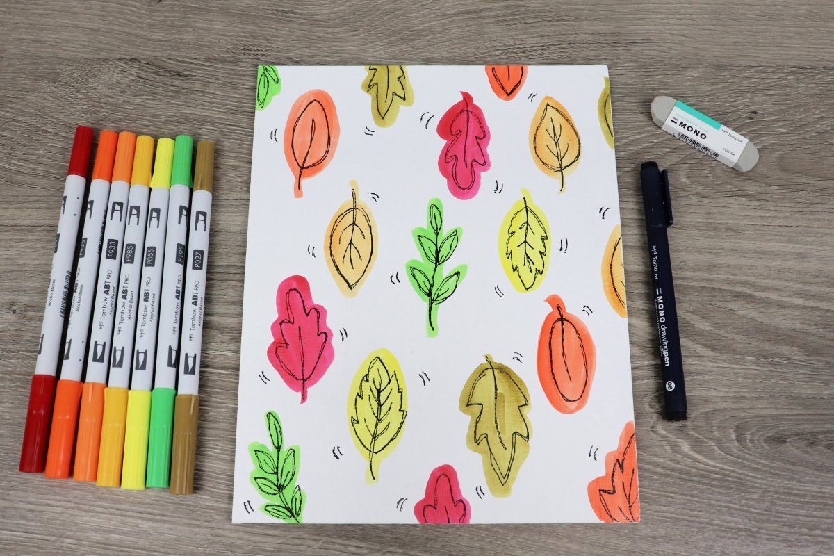 Image contains a canvas decorated with colorful hand drawn fall leaves. Seven markers sit to the left of it, and on the right is a black marker and an eraser.