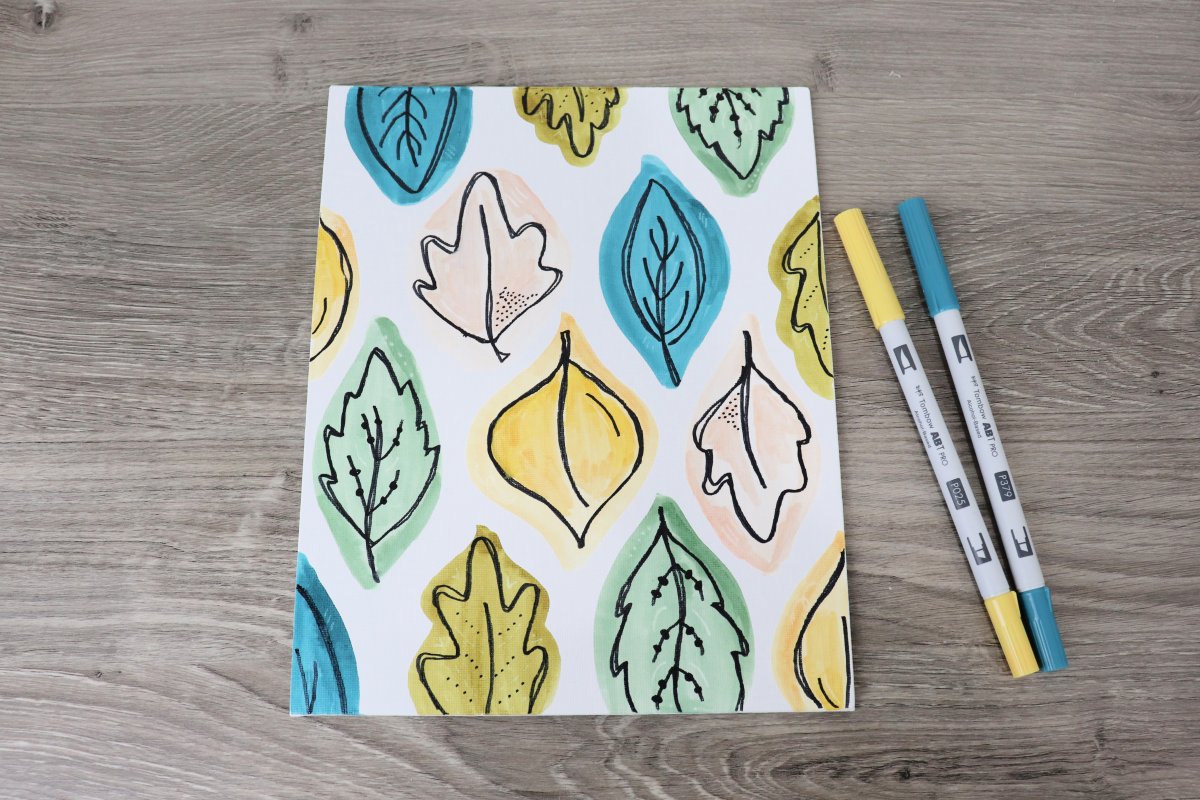 Image contains a white canvas with areas of teal, blush, gold, green, and green-brown and hand drawn leaves. Two markers sit next to the canvas on a wood background.