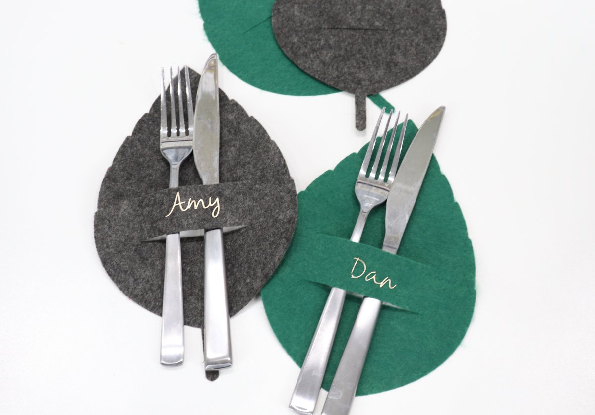 image contains four leaves cut from felt; grey and green. Two of them are labeled with names in gold vinyl and have silverware tucked in the slit.