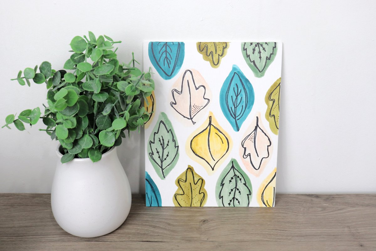 Image contains a white canvas decorated with colorful hand-drawn leaves, next to a pot with faux eucalyptus.