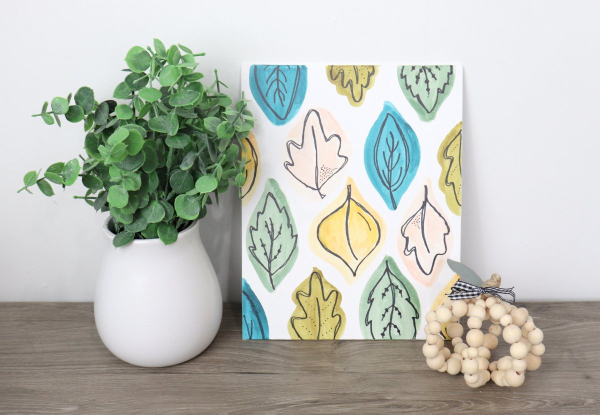 Image contains a canvas with colorful hand-drawn leaves. Next to it is a vase with faux eucalyptus and a wooden bead pumpkin.