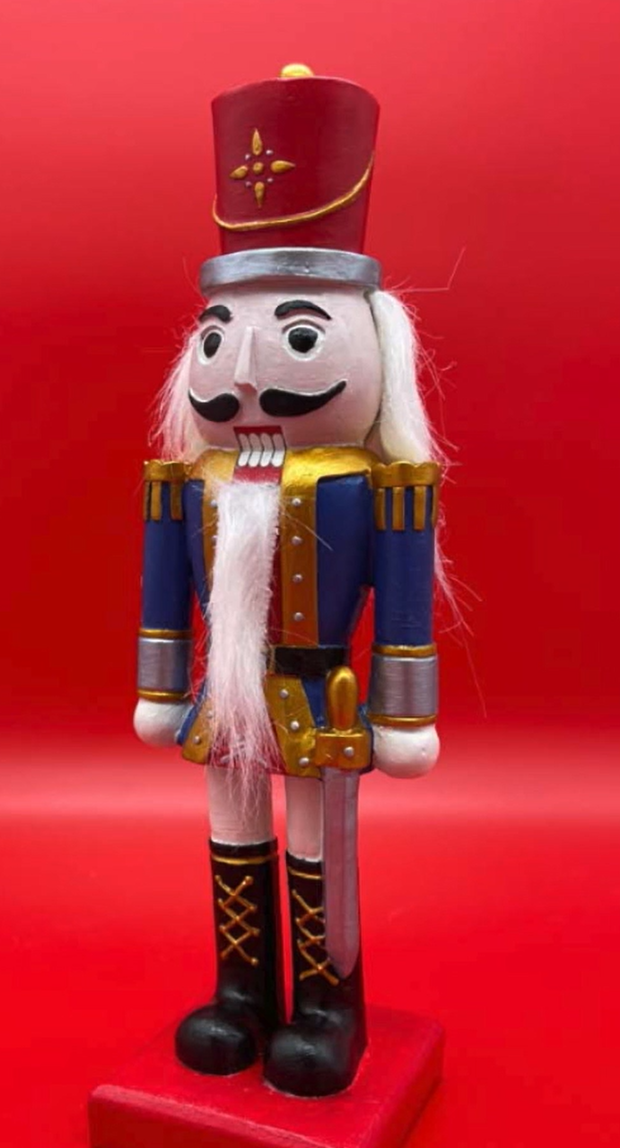 Image contains a hand-painted Nutcracker figure with a blue and gold jacket, black mustache, black boots, and red and gold hat, on a red background.