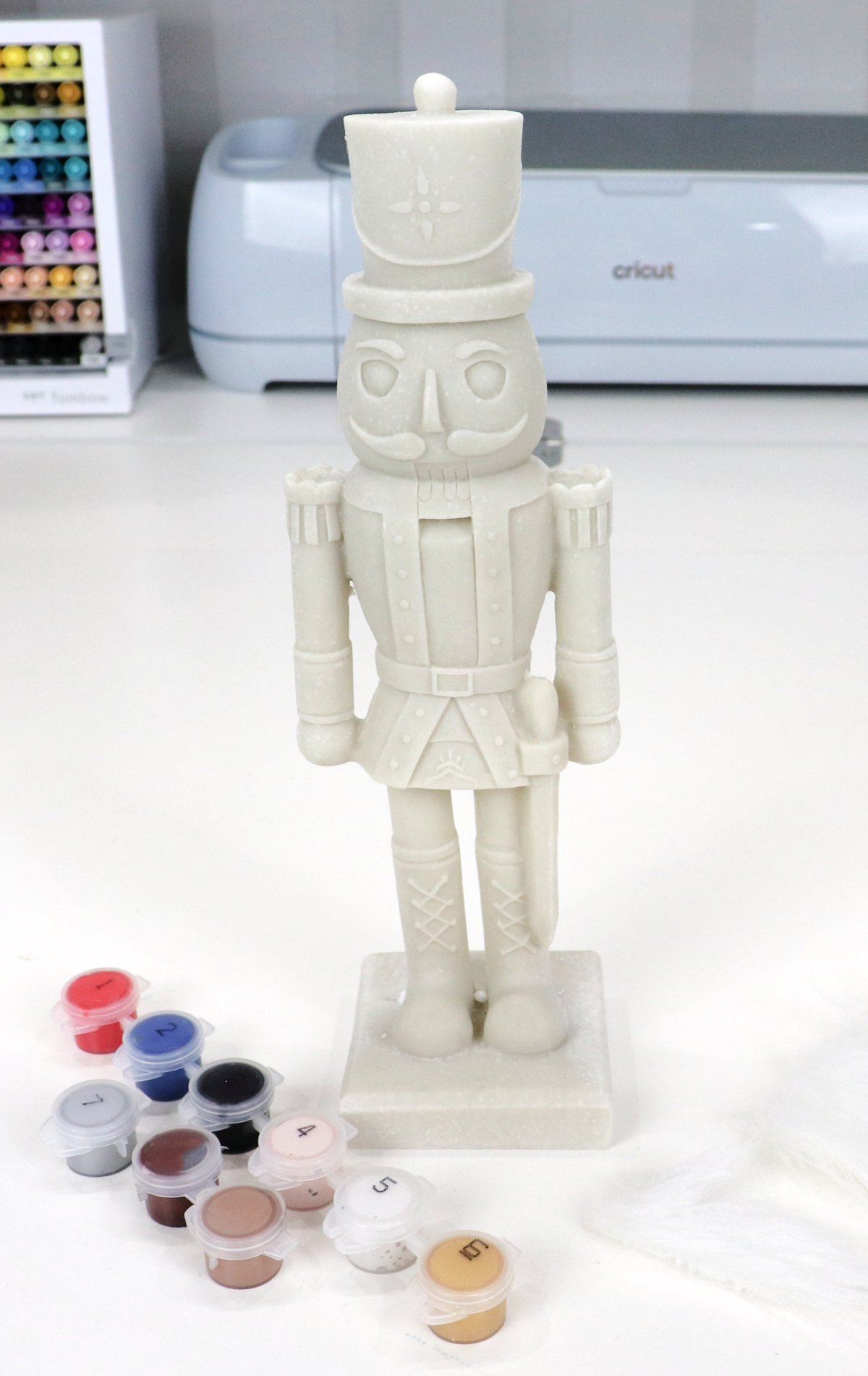 Image contains an unpainted nutcracker figurine and nine colors of paints in small tubs.