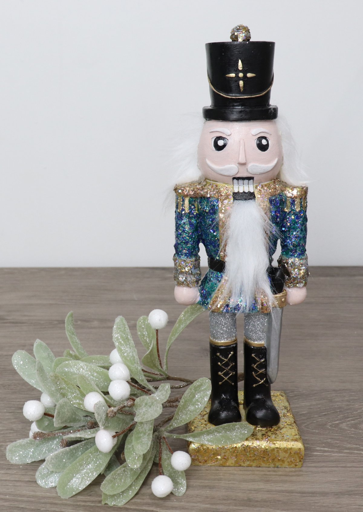 Image contains a hand-painted Nutcracker figurine with a black hat and a glittered blue and gold jacket, next to a sprig of mistletoe.