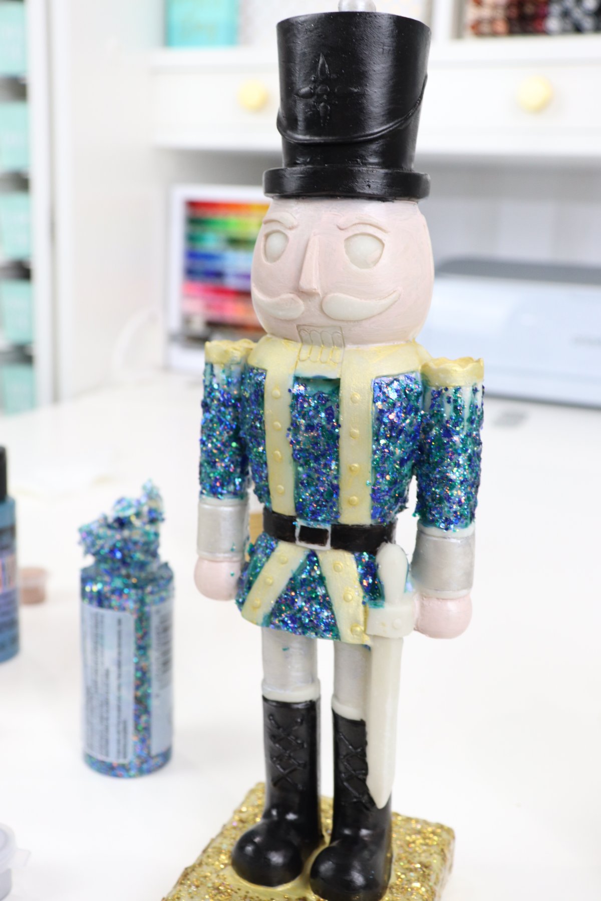Image contains a partially painted and glittered nutcracker figurine with a jar of blue glitter paint beside it.