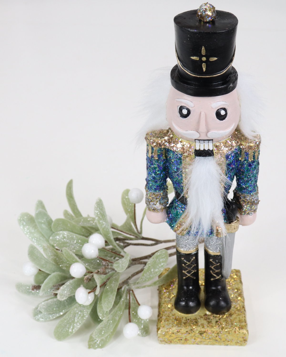 Image contains a handpainted Nutcracker figurine with a black hat and a glittered blue and gold jacket, next to a sprig of mistletoe.