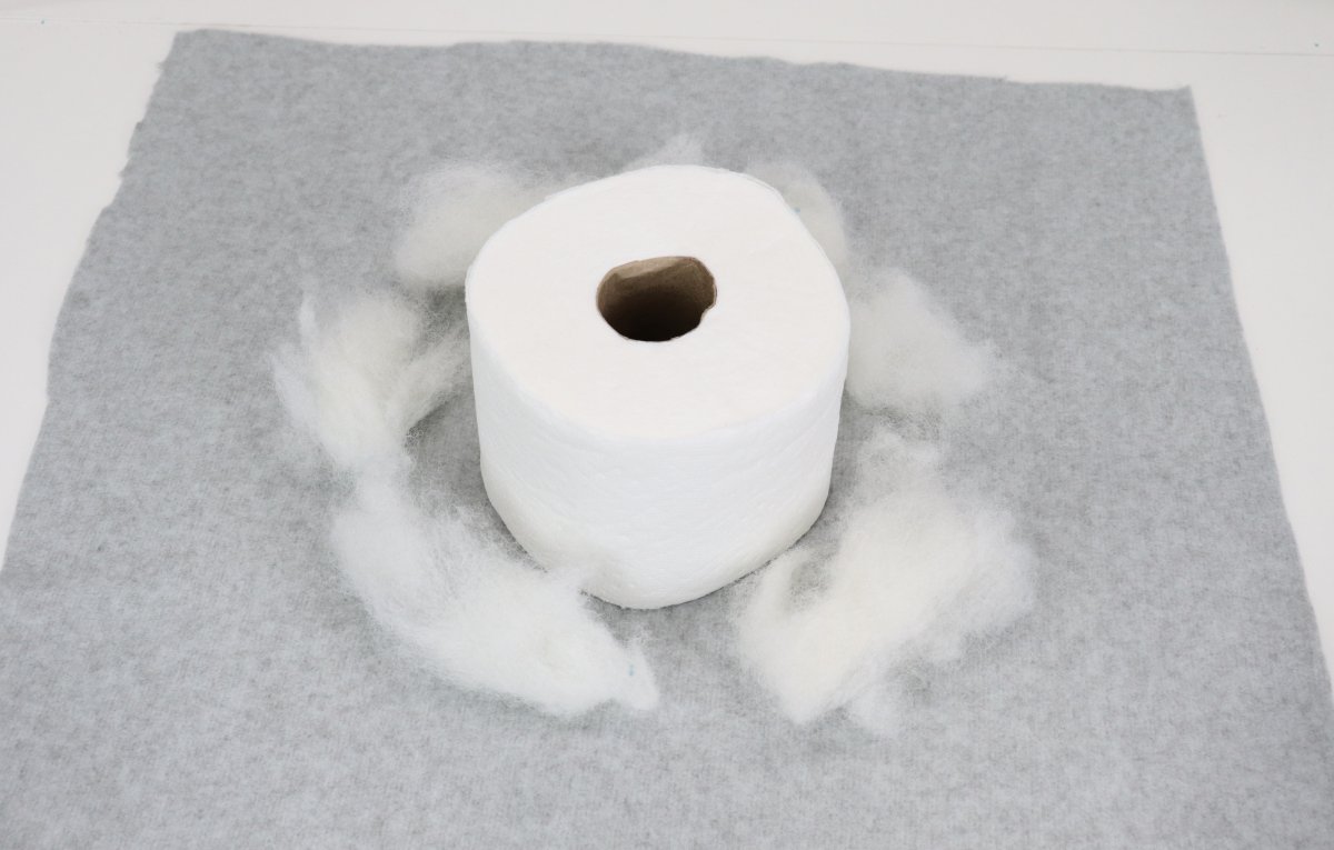 Image contains a roll of toilet paper in the center of a square of grey fleece, surrounded by polyfil.