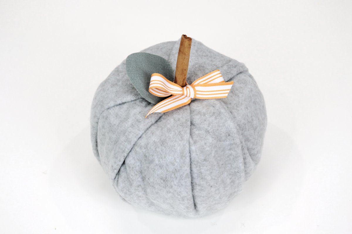 Image contains a pumpkin made of grey fleece, with a cinnamon stick stem, green faux leaf, and orange and white bow.