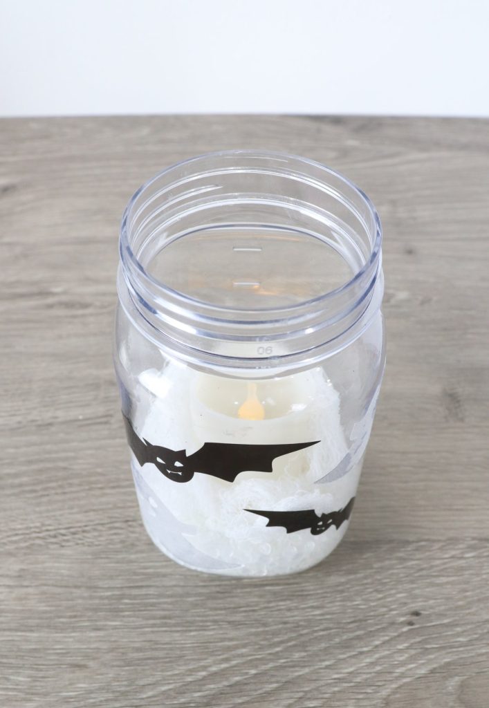 Image contains a mason jar with black bats and white ghosts decorating the outside. Inside, there is gauze and a battery operated candle.