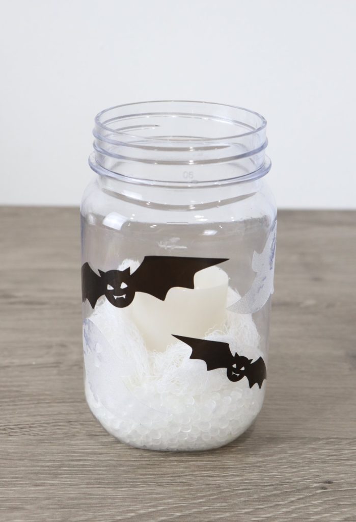 Image contains a mason jar filled with poly pellets, gauze, and a battery operated votive. The outside is decorated with black and white window clings shaped like bats and ghosts.