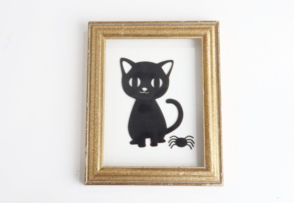 Image contains a gold frame with floating glass, on which there are window clings of a black cat and a spider.