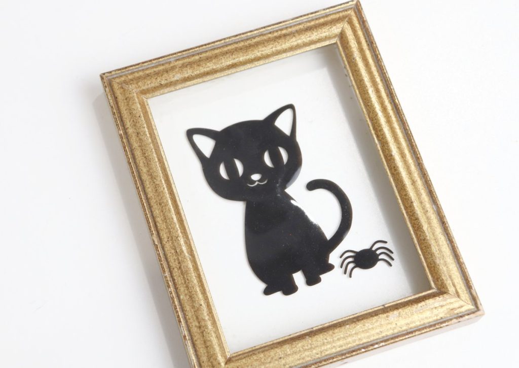 Image contains a small gold frame with floating glass, on which there is a black cat and spider window cling.