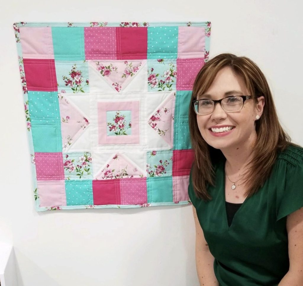 Image contains Amy, smiling, next to a teal and pink mini quilt hanging on the wall.