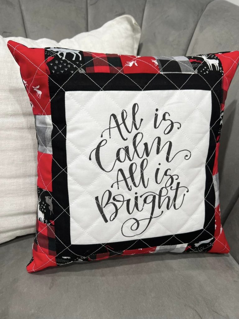Image contains a red, white, black, and grey quilted pillow. In the center is a white panel with the words, "all is calm, all is bright," surrounded by a black frame and red, black, grey, and white patchwork fabric.
