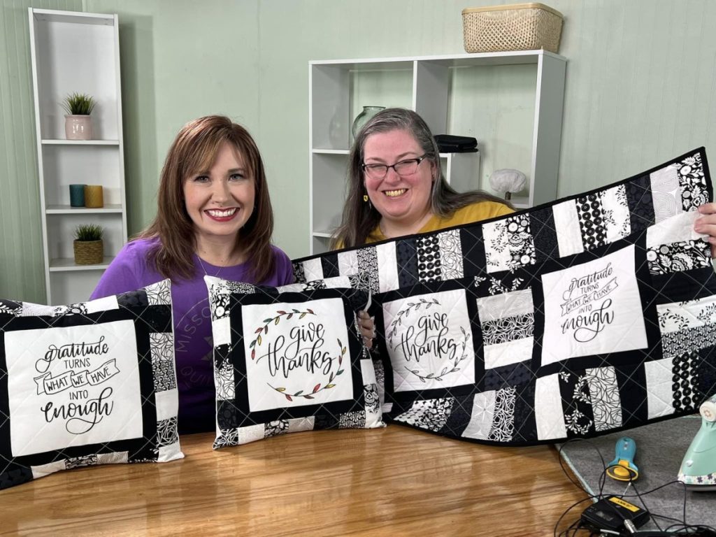 Image contains Amy and Liz holding up two quilted pillows and a quilted table runner.