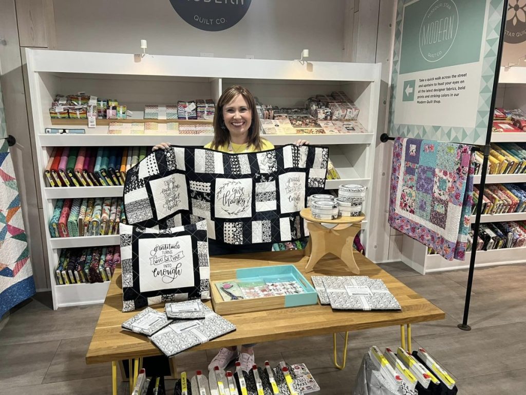 Image contains Amy holding a black and white quilted table runner in the Main Shop of Missouri Star Quilt Company in Hamilton, MO. There are shelves with bolts of fabric behind her, and a table holding a quilted pillow, fabric, and other quilting notions in front.