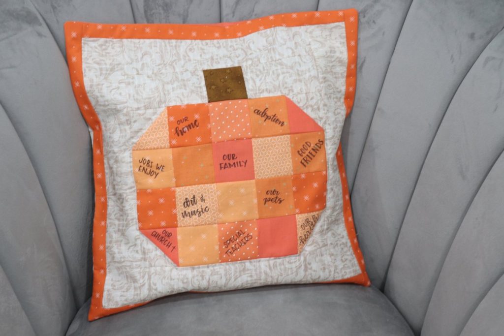 Image contains the pumpkin quilt block turned into a pillow sitting on a grey chair.