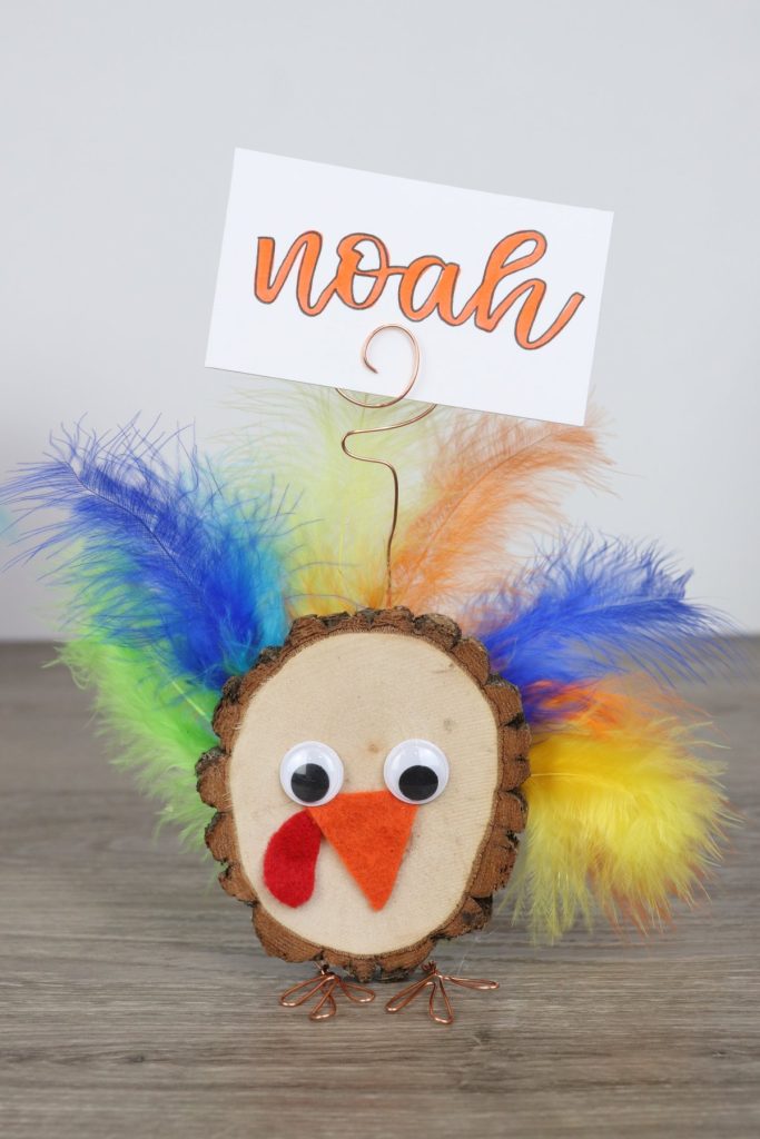 Image contains a wood slice turkey made with feathers, google eyes, felt, and wire. It has a place card attached with the name "Noah" written on it in orange marker.