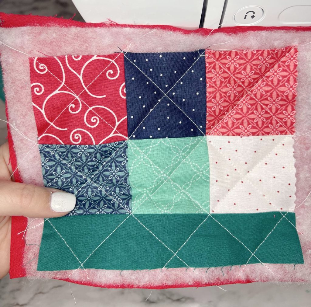 Image contains Amy's hand holding a quilt sandwich with the patchwork piece on top, then batting and the lining.