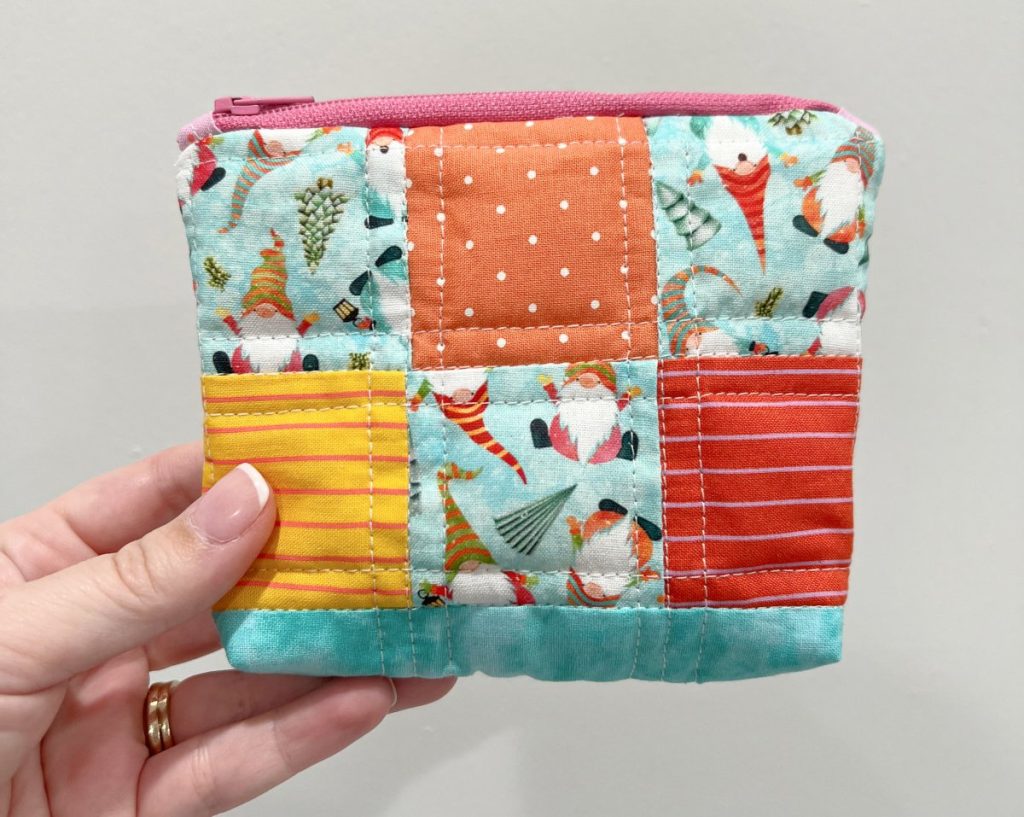 Image contains Amy's hand holding a zipper pouch made of teal, orange, yellow, and red fabric with gnomes.