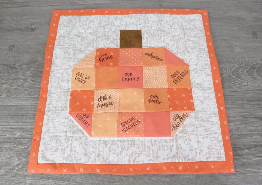 Image contains a beige and orange quilt block that features a patchwork pumpkin. Words are written on the pumpkin with black fabric marker.